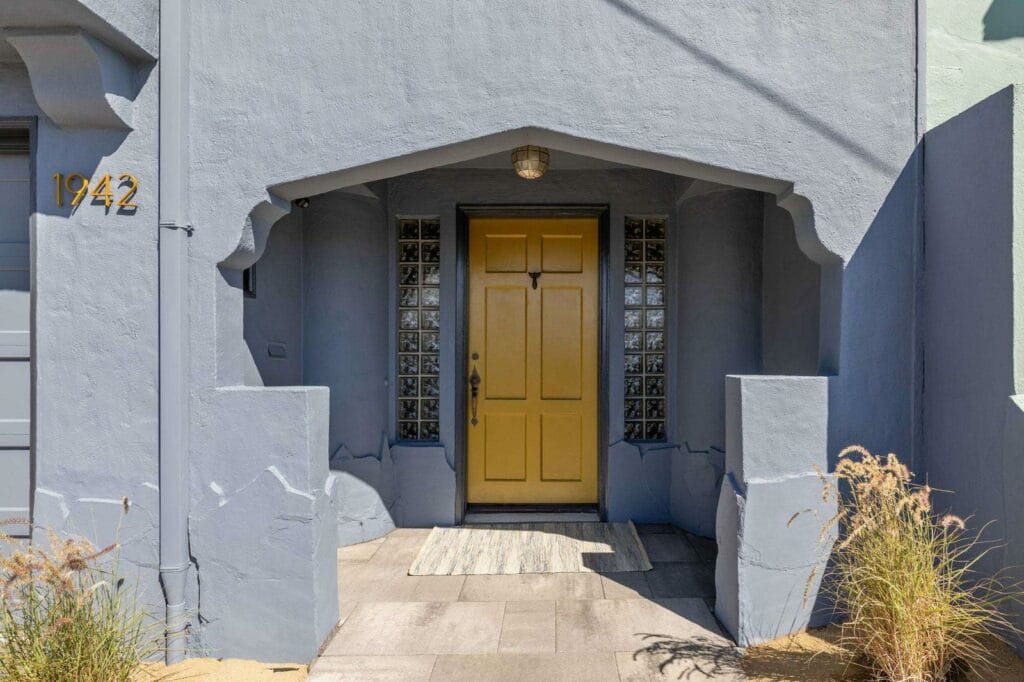 A yellow front door with glass block windows set in a gray stucco entryway, house number 1942.