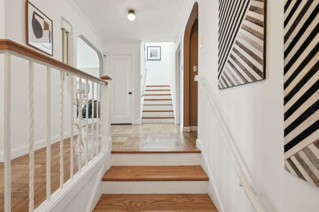 Bright hallway with wooden stairs, white walls, framed art, and a decorative railing on the left.